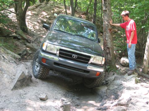 1990 Ford Bronco - Toyota Tacoma tackling a challenging off-road course in a wooded area.