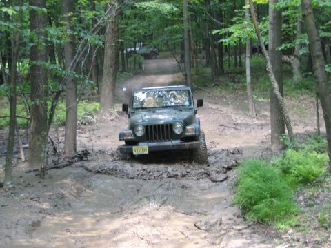 1990 Ford Bronco - Jeep Wrangler tackling a challenging muddy off-road course in a forest setting.