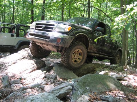 1990 Ford Bronco - A black Toyota Tacoma is shown off-roading over rocks with a white Jeep in the background.