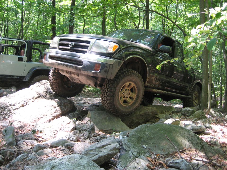 Black Toyota Tacoma navigating rocks on a wooded trail, white Jeep visible to the left