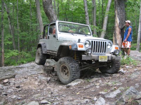 1990 Ford Bronco - A silver Jeep Wrangler navigates a rocky trail in a wooded area.