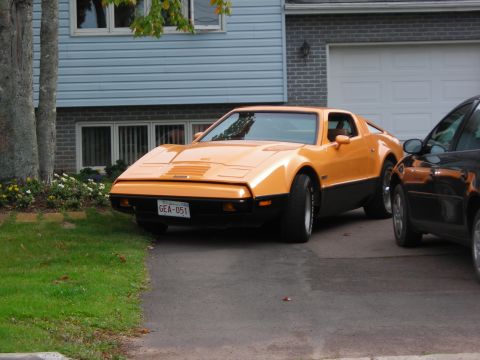 1984 Ford Bronco - This orange sports car, identified by its license plate GEA-051, is parked in a driveway next to another vehicle.