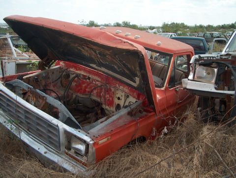 1979 Ford Bronco - This orange pickup truck, likely from the mid-1970s, sits in a junkyard with its hood ajar.