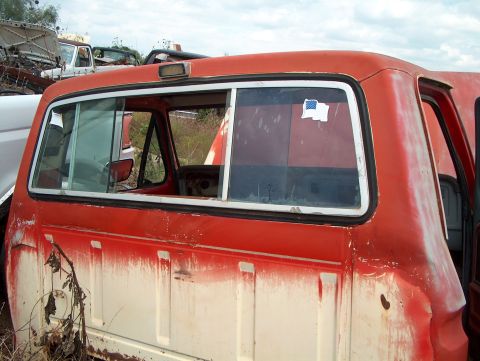 1979 Ford Bronco - A weathered red and white pickup truck sits in a junkyard with other vehicles in the background.