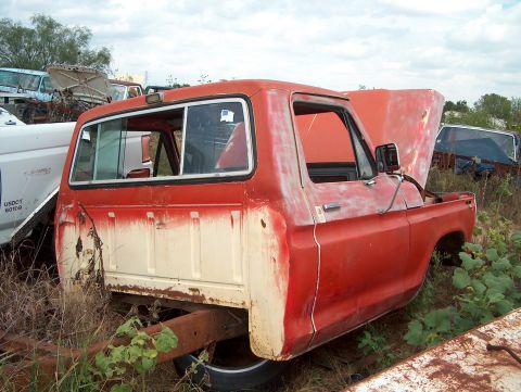 1979 Ford Bronco - This abandoned truck cab sits in a junkyard, surrounded by other vehicles under an overcast sky.