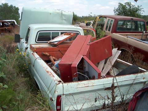1979 Ford Bronco - A light blue pickup truck sits in a junkyard, its bed filled with debris and other vehicles visible vehicles.