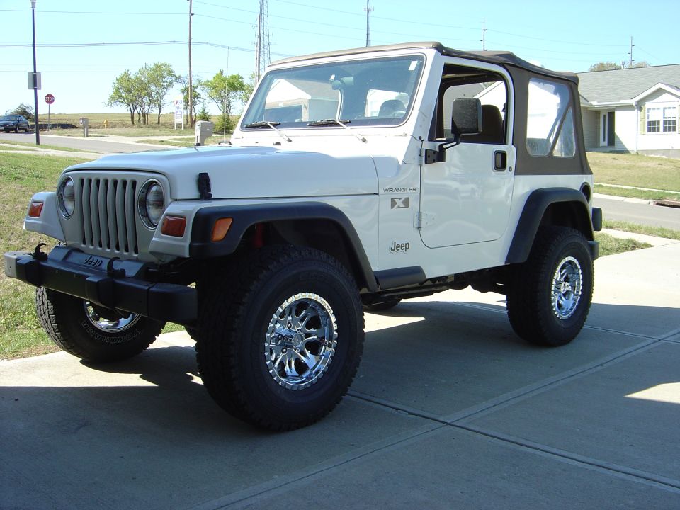 White Jeep Wrangler X with soft top and chrome wheels on oversized tires, parked in a driveway, front three-quarter view