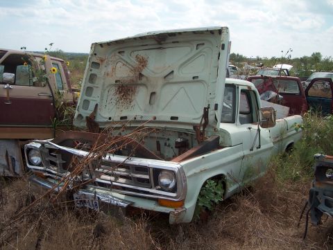 1979 Ford Bronco - This old pickup truck sits abandoned in a junkyard, overgrown with weeds and debris.