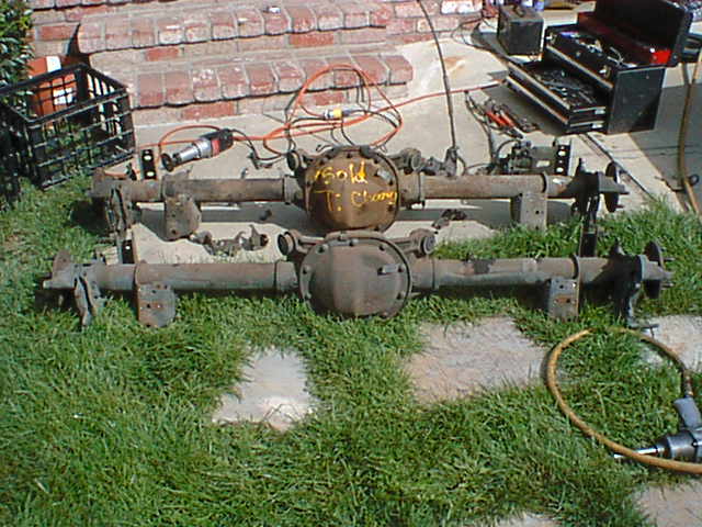 Two rusty solid rear axle assemblies on grass; one diff cover marked SOLD in yellow