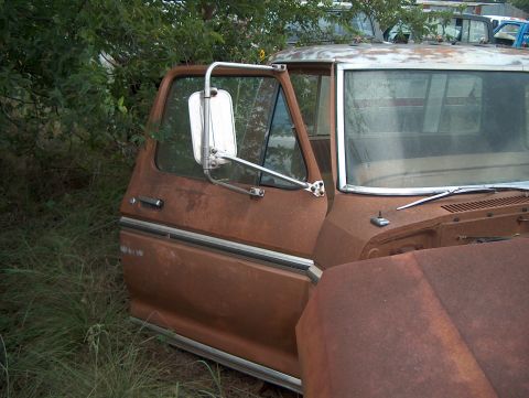 1979 Ford Bronco - A weathered pickup truck sits in a junkyard, its door ajar, surrounded by overgrown grass and other vehicles.