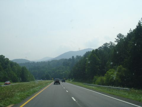 1984 Ford Bronco - Multiple vehicles drive along a highway surrounded by lush green trees and distant mountains under a hazy sky.