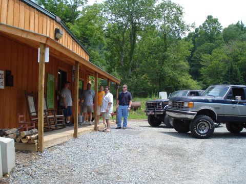 1984 Ford Bronco - Two lifted Ford trucks are parked in front of a rustic building. Several men are standing on the porch.