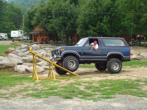 1984 Ford Bronco - A blue Ford Bronco navigates an off-road obstacle course, with its front tires climbing over a yellow ramp and rocks.