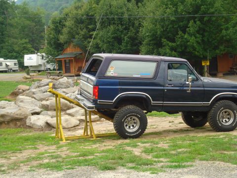 1984 Ford Bronco - A dark blue SUV with a purple top is positioned on a yellow ramp in a grassy area with trees and buildings in the background.