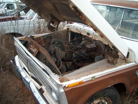 1979 Ford Bronco - Engine bay of a 1979 Ford Bronco in a junkyard, with another vehicle partially visible.