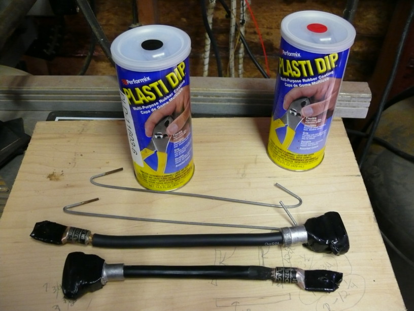 Two cans of Performix Plasti Dip and two automotive parts with black coating and metal fittings on a wooden surface