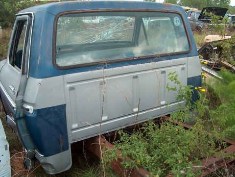 1979 Ford Bronco - The rear of a 1979 Ford Bronco shows its two-tone blue and silver paint scheme and a cracked rear window, surrounded by overgrown weeds.