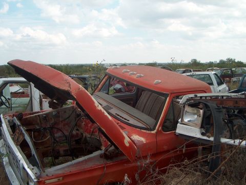 1979 Ford Bronco - Junkyard scene with an abandoned orange pickup truck and other vehicles under a cloudy sky.