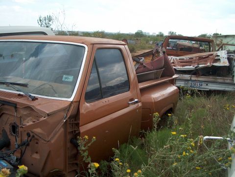 1979 Ford Bronco - A rusty brown pickup truck sits in a junkyard, with other vehicles visible in the background.
