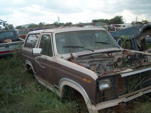 1979 Ford Bronco - A weathered Ford Bronco sits abandoned in a junkyard, its engine compartment exposed and vegetation encroaching.