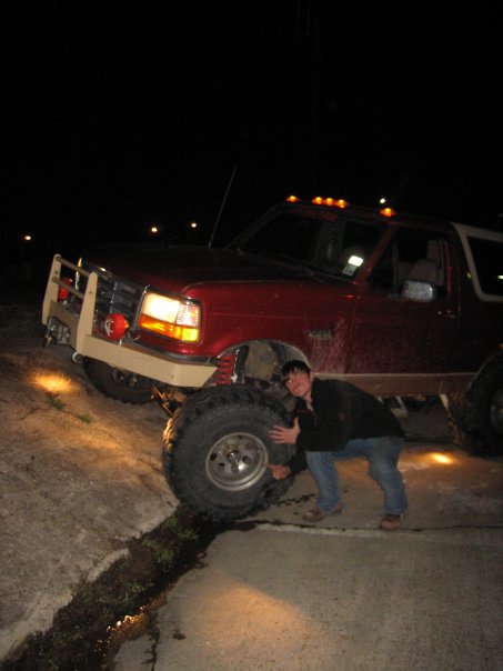 1995 Ford Bronco - A red Ford Bronco with oversized tires and a custom bumper is shown at night with a person next to it.