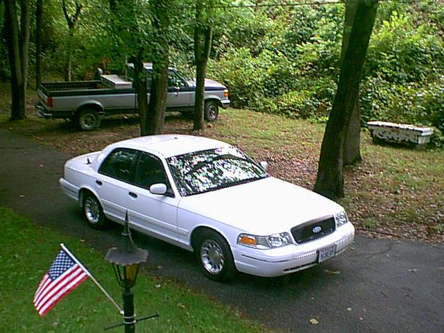 White Ford sedan parked in driveway with a pickup truck behind it and trees around.