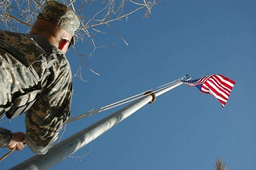 Soldier in camouflage uniform pulls rope to raise American flag on flagpole against clear blue sky