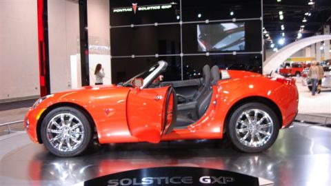 1996 Ford Probe - Orange Pontiac Solstice GXP convertible with doors open, chrome wheels, on display at an auto show.