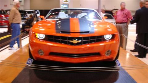 1996 Ford Probe - An orange Chevrolet Camaro convertible is displayed at an auto show with people in the background.
