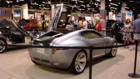 1996 Ford Probe - A silver concept car with its gullwing doors open is displayed at an auto show with other vehicles and people.