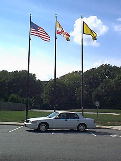 1998 Ford Crown Victoria - A white 1998 Ford Crown Victoria is parked in a lot with three flagpoles in the background.