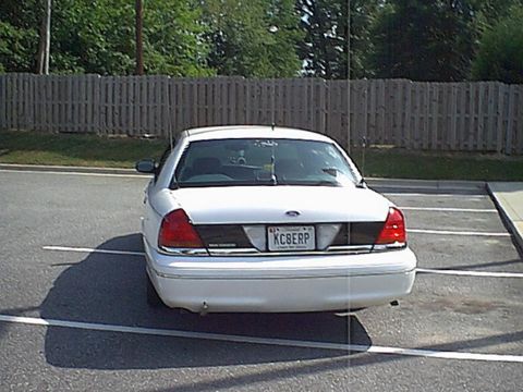 1998 Ford Crown Victoria - Rear view of a white sedan parked in a lot with a wooden fence in the background.