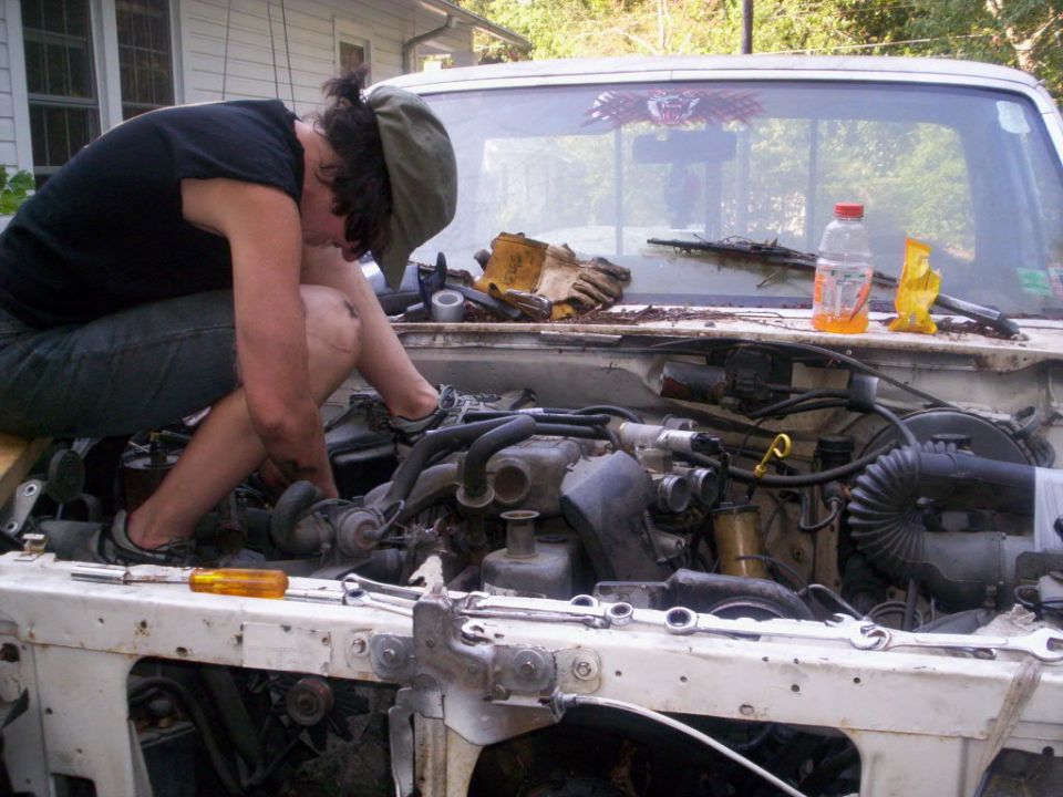 Person crouched inside the open engine bay of a white pickup, working on the engine with tools nearby