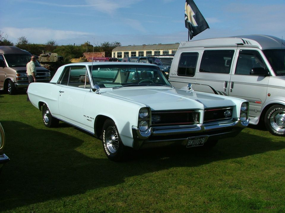White 1965 Pontiac Catalina coupe parked on grass, front 3/4 view, with a van and other vehicles in the background