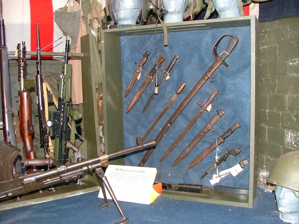 Display of firearms and bayonets, including a machine gun and swords, with a helmet on the right