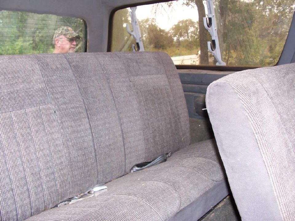 Gray cloth bench seat interior with seat belts; gun rack in rear window and man in camo hat outside