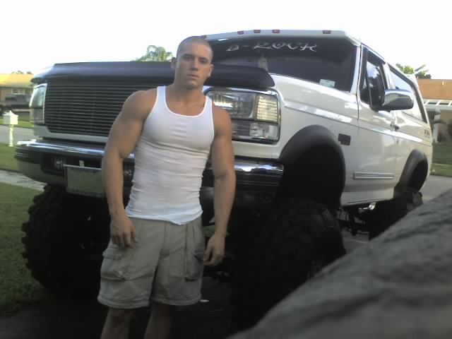 Man in white tank top and khaki shorts stands in front of a lifted white 1992 Ford Bronco with large tires