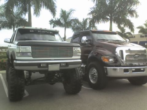 1995 Ford Bronco - Two custom trucks are parked side-by-side in a parking lot with palm trees in the background.