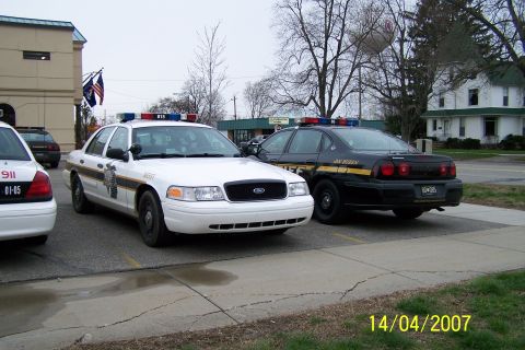2003 Ford Crown Victoria - A white 2003 Ford Crown Victoria police car is parked in front of a building, with a black police car to its right.