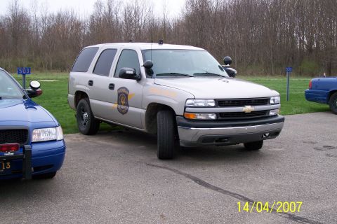 2003 Ford Crown Victoria - Two police vehicles, a silver SUV and a blue sedan, are parked on asphalt with a grassy area and trees in the background.