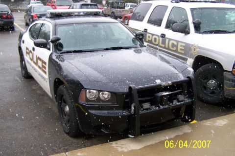 2003 Ford Crown Victoria - A black police car with a push bar is parked next to a white police SUV, with snow falling.
