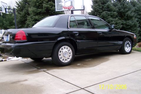 2003 Ford Crown Victoria - A black sedan is parked on a wet driveway with trees and a basketball hoop visible.