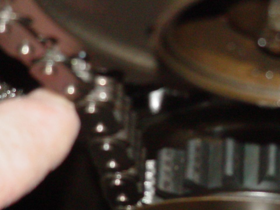 Close-up of a metal timing chain with rivets and a toothed gear, with a finger visible on the left