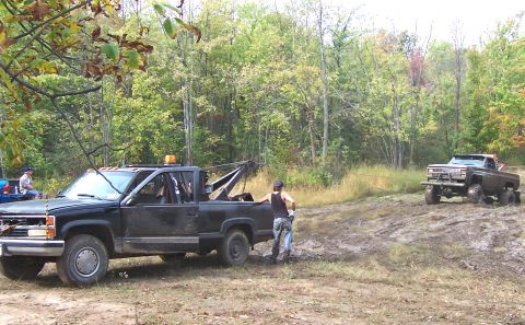1978 Ford Bronco - Two trucks are in a muddy area with trees in the background. One truck is a tow truck, the other is an off-road vehicle.