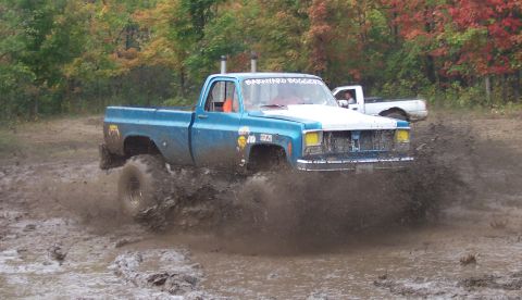 1978 Ford Bronco - Pickup truck powers through a muddy track, kicking up a large spray of mud. Another truck is visible in the background.