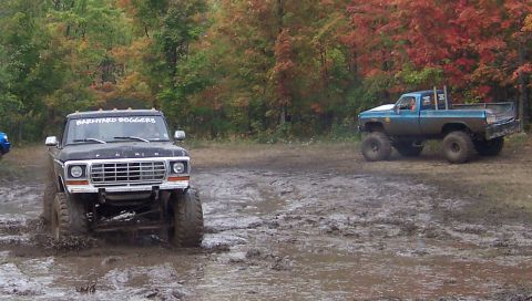 1978 Ford Bronco - Two SUVs navigate a muddy off-road trail during autumn, with colorful foliage in the background.