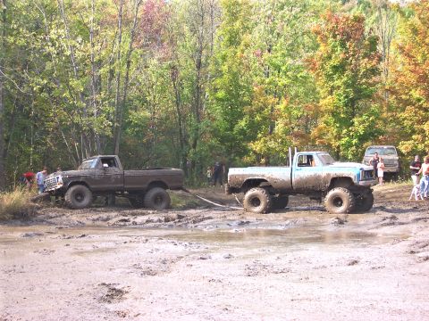 1978 Ford Bronco - Two trucks are stuck in a muddy field with trees in the background, one is being pulled by the other.