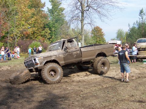 1978 Ford Bronco - Muddy 1977 Ford F-150 truck stuck in a mud pit during an event. Spectators watch from the side.