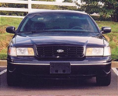 1999 Ford Crown Victoria - A dark blue Ford Crown Victoria is parked in front of a white fence, showing its front grille and headlights.