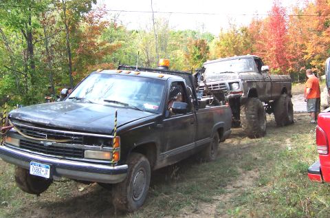 1978 Ford Bronco - A lifted Ford truck is being towed by a Chevrolet pickup truck in a wooded area with fall foliage.